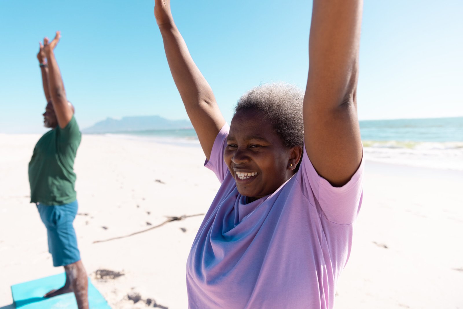 smiling african american senior woman and man raising arms and exercising at beach against clear sky. copy space, unaltered, love, together, yoga, retirement, nature, vacation and active lifestyle.