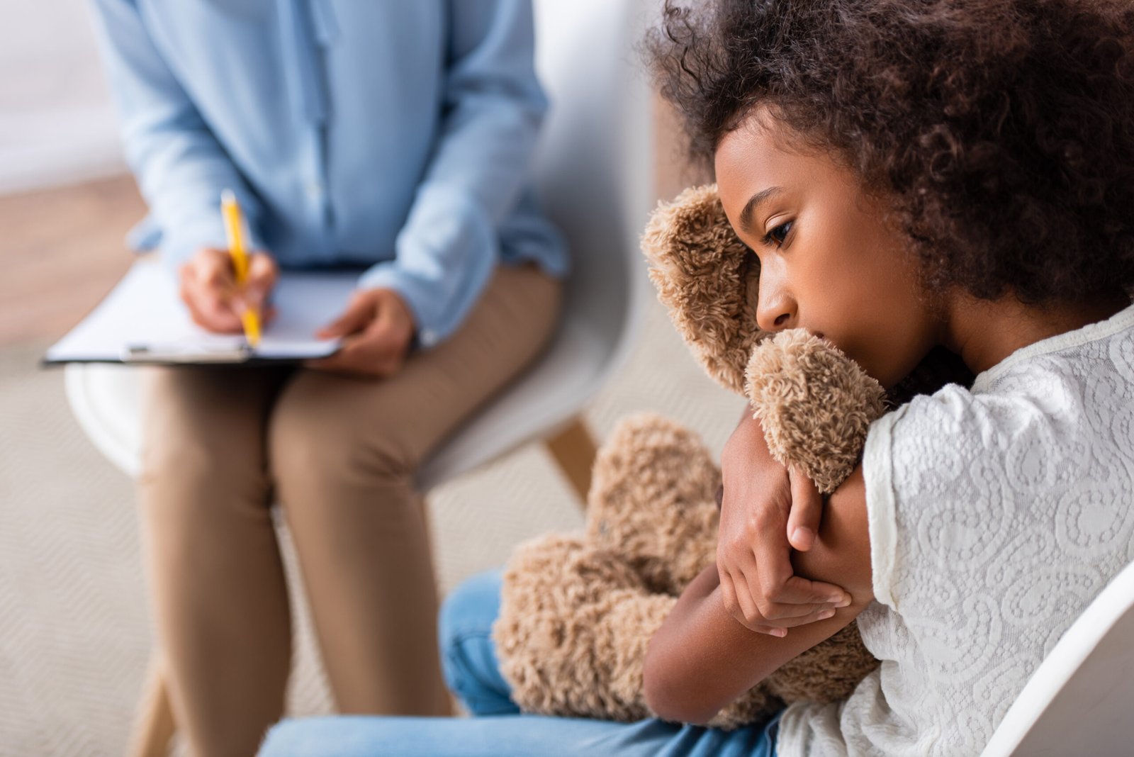upset african american girl with autism hugging teddy bear during consultation with blurred